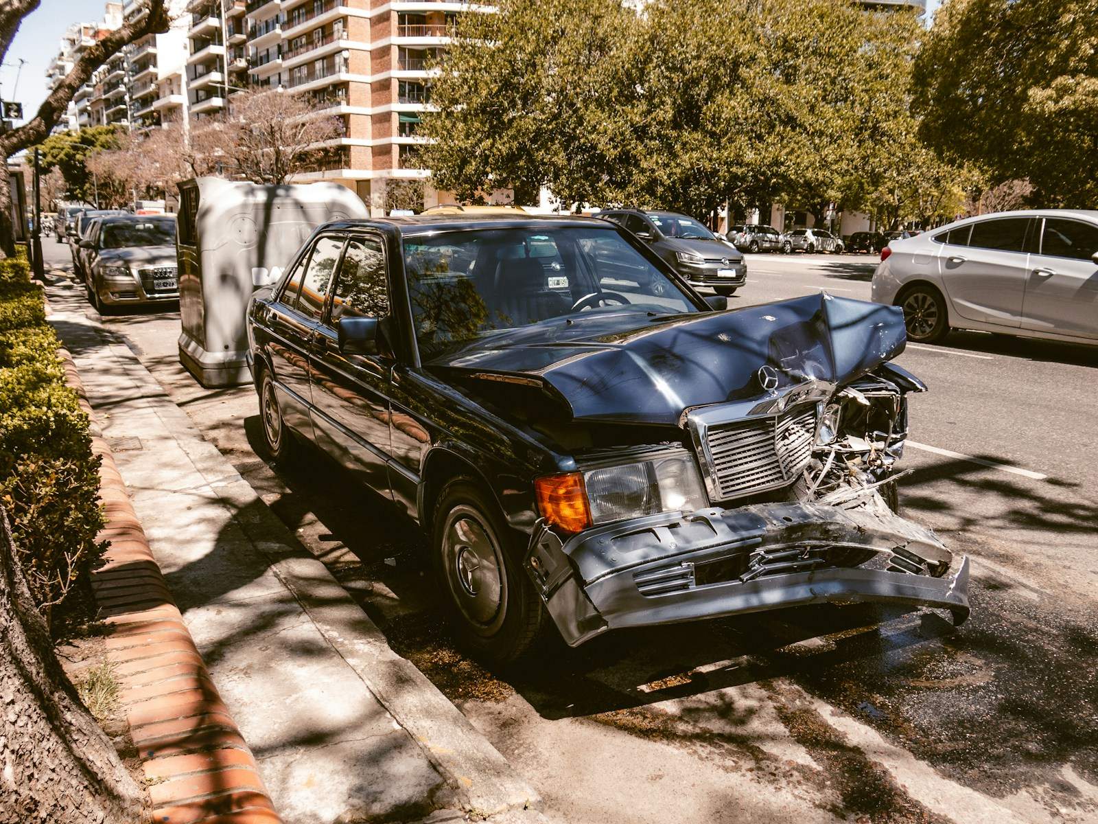 Czarny sedan z rozbitym przodem zaparkowany przy ulicy po kolizji, przed naprawą